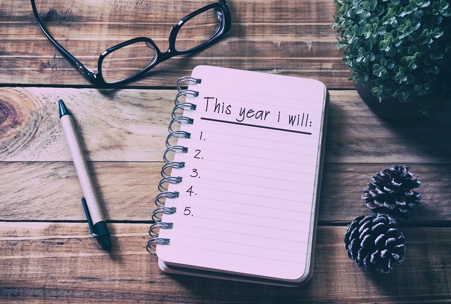 Notebook on wooden table surrounded by a pen, pinecones, classes, and a plant. The notebook says "This year I will" and is followed by a numbered list.