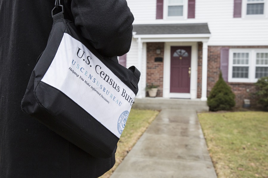 Close-up of a messenger bag with U.S. Census Bureau information on the outside. The person wearing the bag is approaching a house.