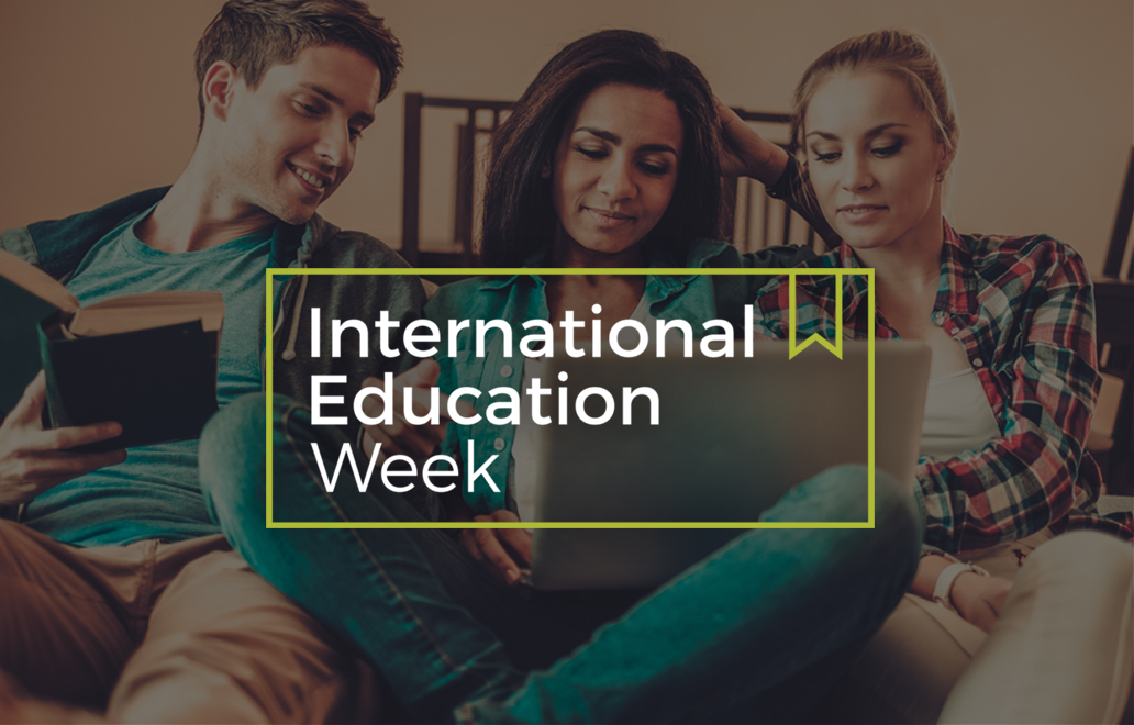 Three people sitting together looking at a person's laptop, with a banner saying "International Education Week" over top