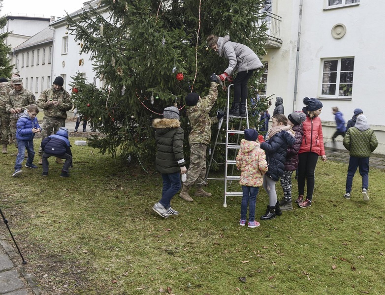Children and soldiers gathered around a pine tree. A soldier is handing decorations to a woman on a ladder