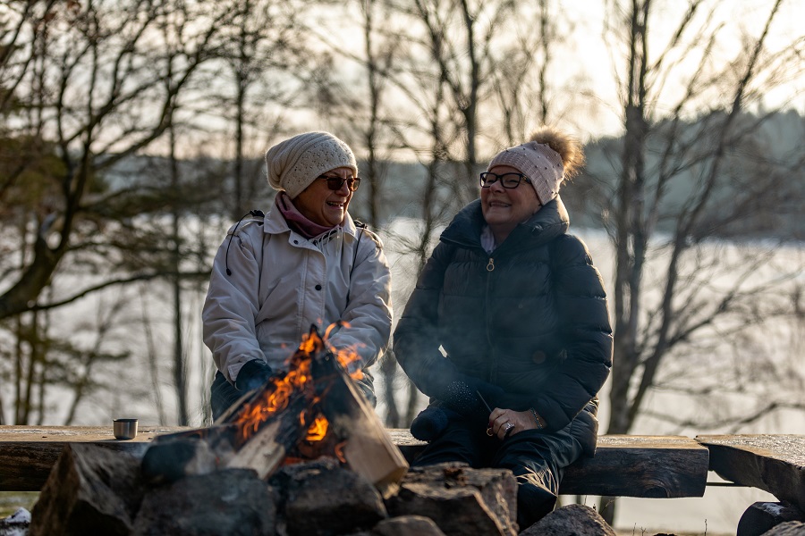 Two senior ladies are sitting on a bench by a burning campfire​ during wintertime.
