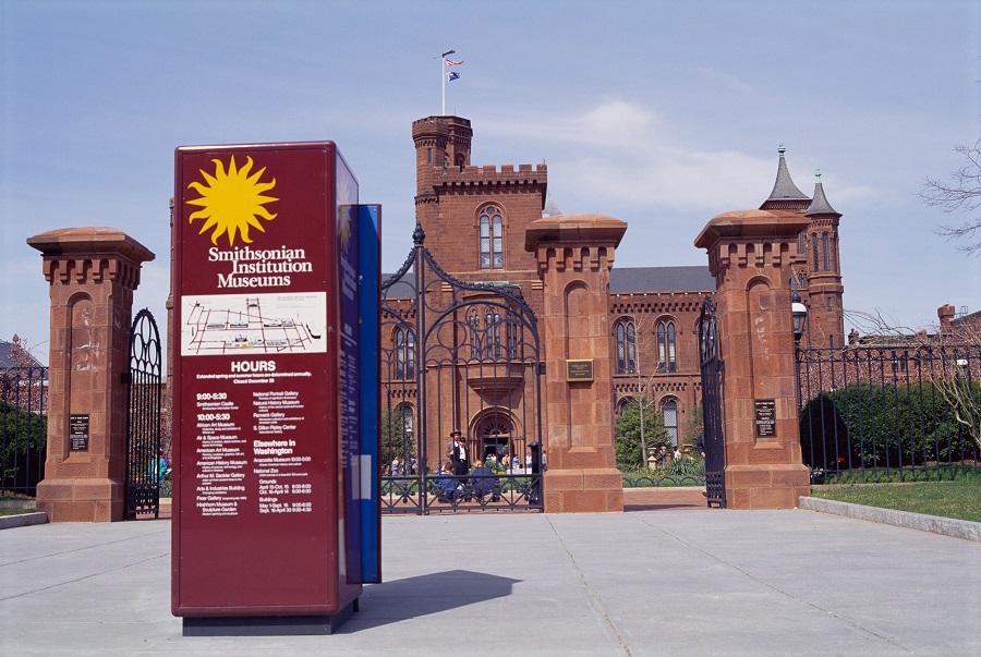 Image of the Smithsonian Castle, with a pillar in the foreground showing Smithsonian building hours