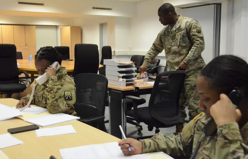 Two U.S. Army Team Trace members sit at a table, talking on the phone and writing notes. One person stands and supervises.