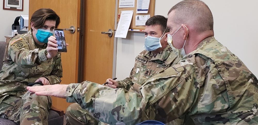 Three men in Army uniforms sit together, all wearing face masks. One is holding up a cell phone with a Facetime call in progress.