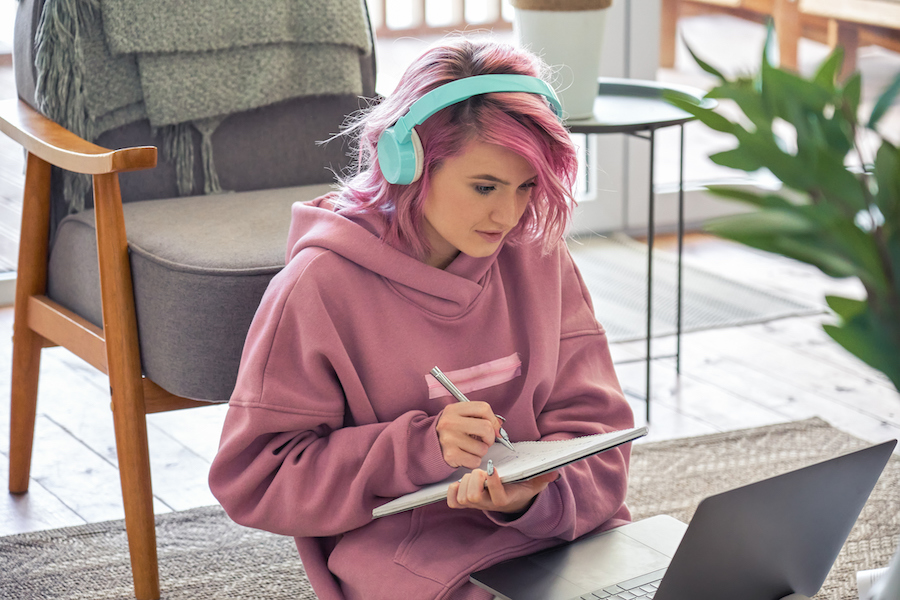A woman with pink hair, wearing a pink hoodie and light blue headphones, is sitting on the floor with her back to a chair and a laptop on her lap. She is holding a notepad and is writing on it.