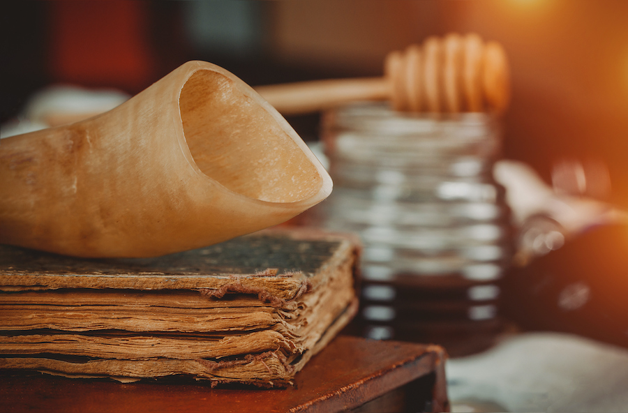 A close-up shot of a shofar, or ram's horn, sitting on top of a worn book. In the background is a jar of honey with a wooden honey dipper on top.