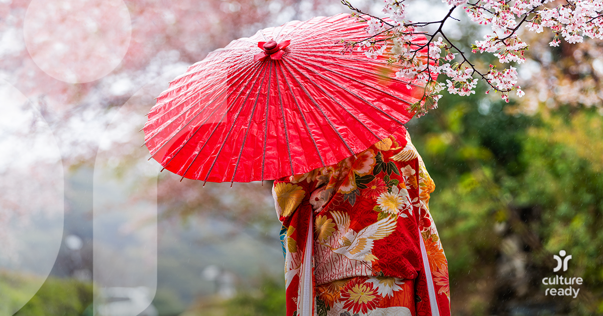 A woman in a red kimono holds a red umbrella in front of cherry blossom flowers