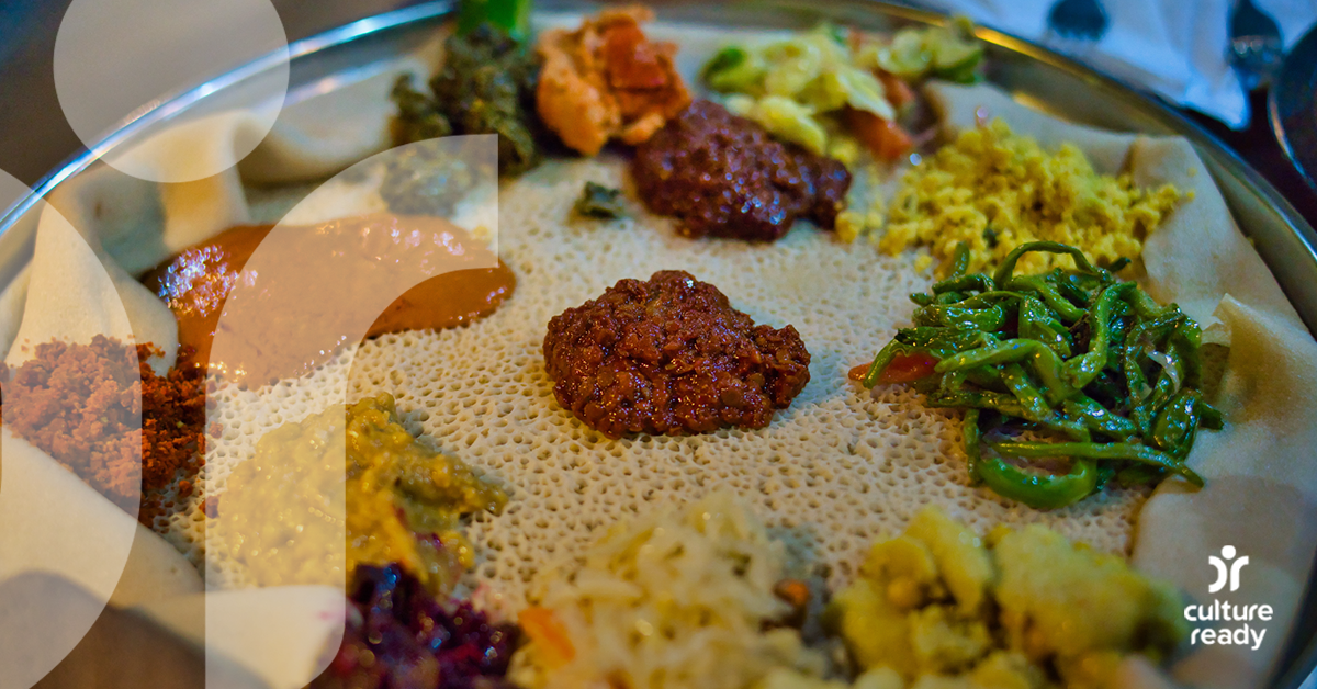Injera bread laid flat becomes a beyaynetu platter with colorful Ethiopian dishes in yellows, oranges, and green