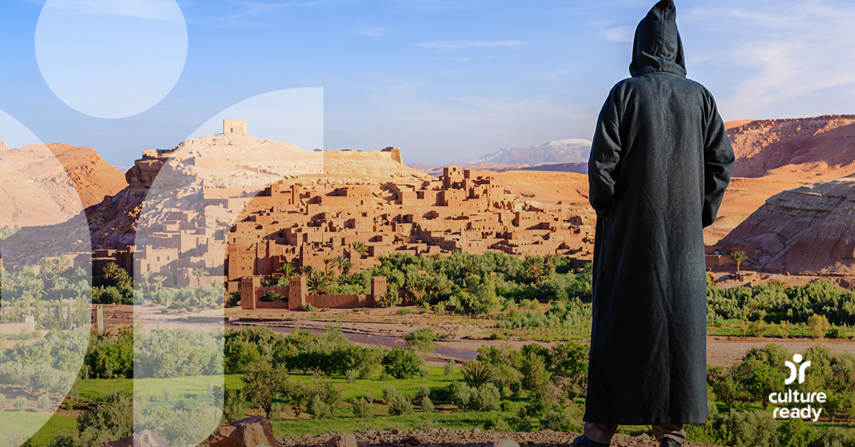 A person wearing a djellaba, a long black tunic with a pointed hood, looks off into the distance at a blue sky, light tan mountains, and a green foreground.