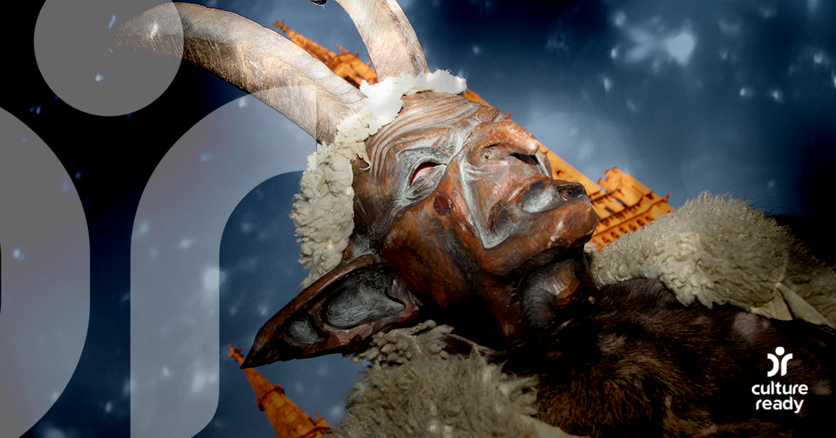 A man wearing a carved wooden mask and animal horns depicting Krampus stands in front of a church with a night sky overhead