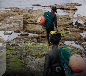 Women in diving suits walking on a beach, carrying fishing nets.
