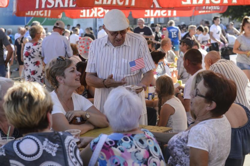 An older man holding an American flag stands talking to a group of people sitting at a table at a Fourth of July/Independence Day cookout in Miroslawiec, Poland.