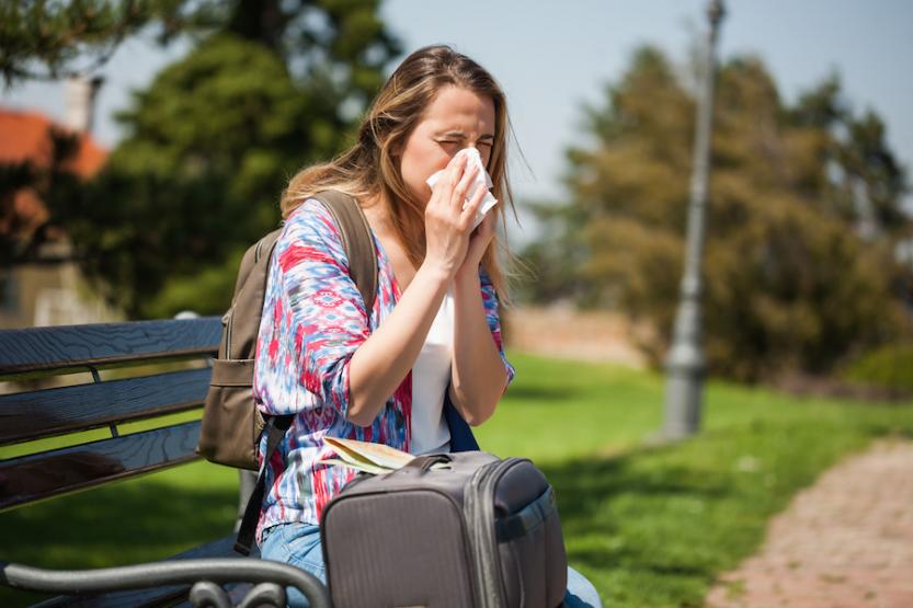 Woman wearing a backpack sitting on a bench, sneezing into a tissue next to a suitcase with a map on top.