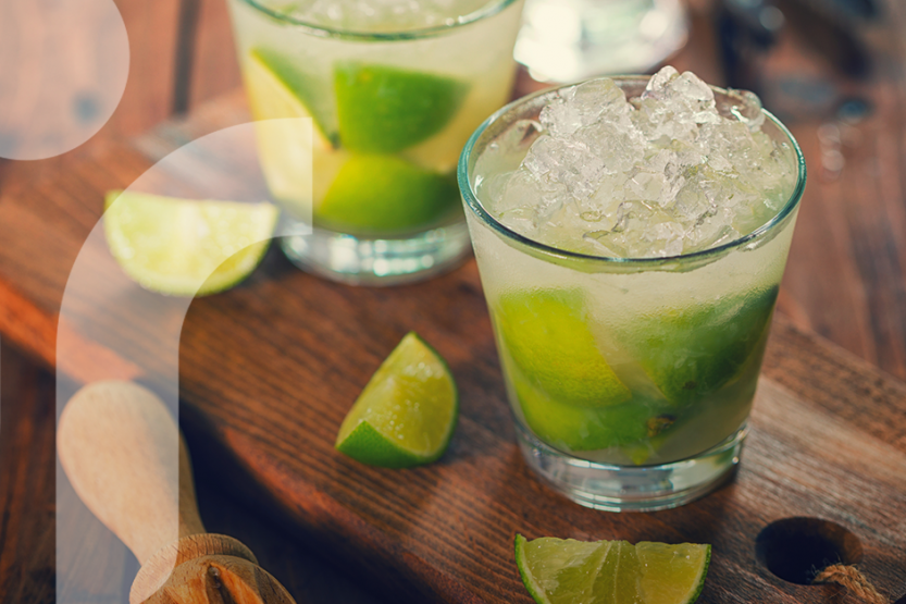 Two cocktail glasses filled with ice, cachaca, and limes sit on a wooden counter with three more slices of lime