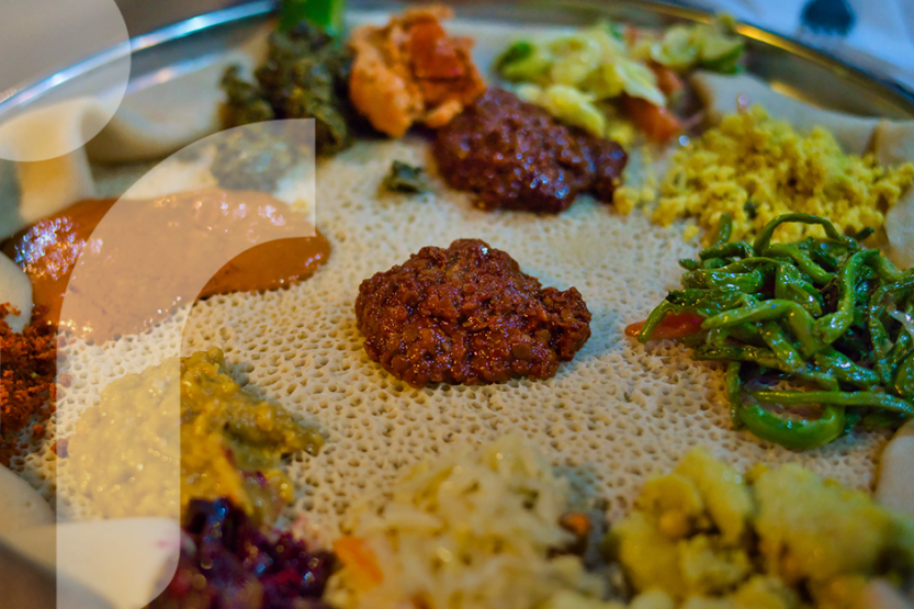 Injera bread laid flat becomes a beyaynetu platter with colorful Ethiopian dishes in yellows, oranges, and green