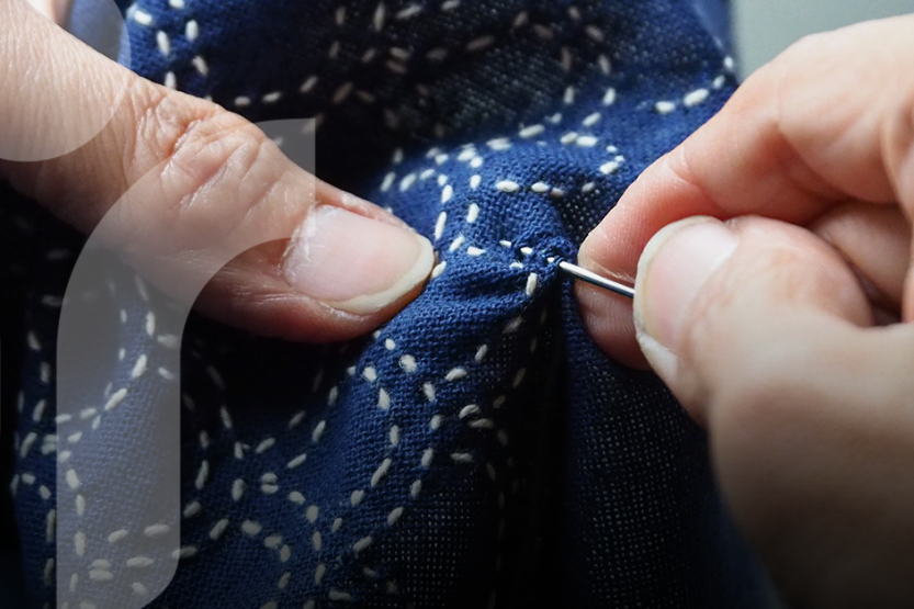 A pair of hands use a needle to thread a pattern with white thread into blue fabric