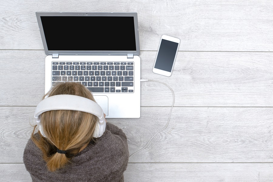Girl wearing headphones lying on the ground with a laptop open in front of her and a smartphone next to the laptop
