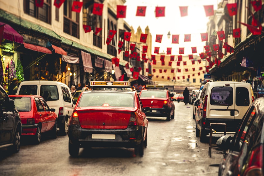 Red taxis in Fez, Morocco, driving down a street with cars parked on either side. Banners of the Moroccan flag hang between buildings.