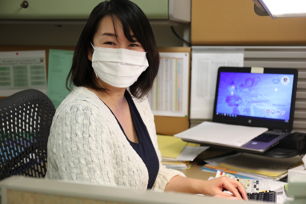 A woman wearing a face mask poses for a photo, with a laptop on a desk behind her.