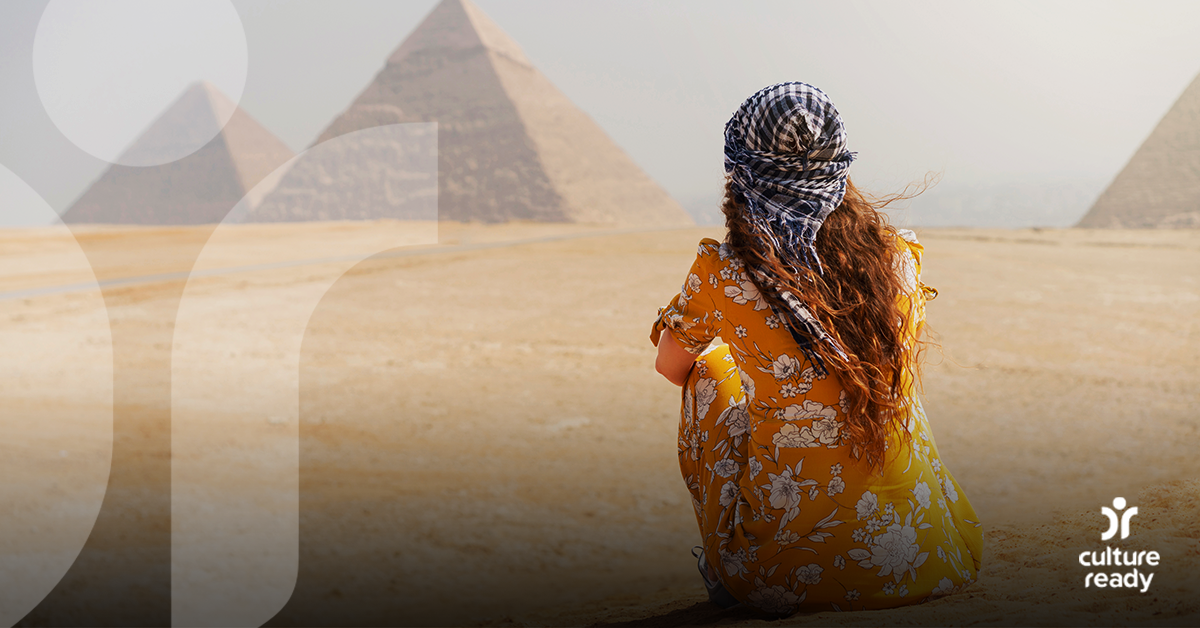 A woman wearing a bright orange dress and a blue checked headscarf facing away from the camera. She is looking at Egyptian pyramids in the distance.