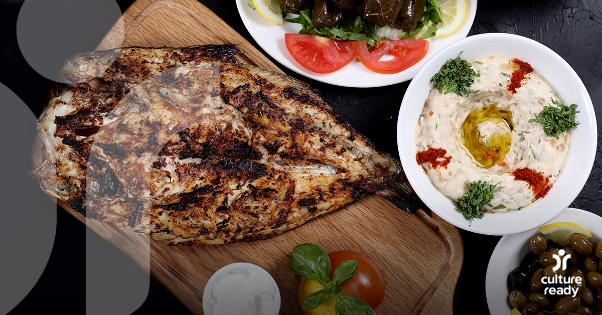 A large piece of grilled fish on a cutting board with vegetables, hummus, and olives on the side