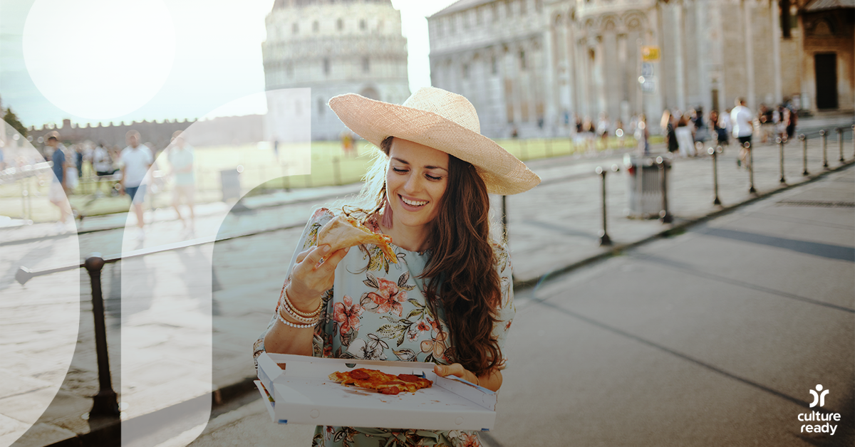 A woman in a floral dress and straw hat eats a slice of pizza in front of a Roman cityscape