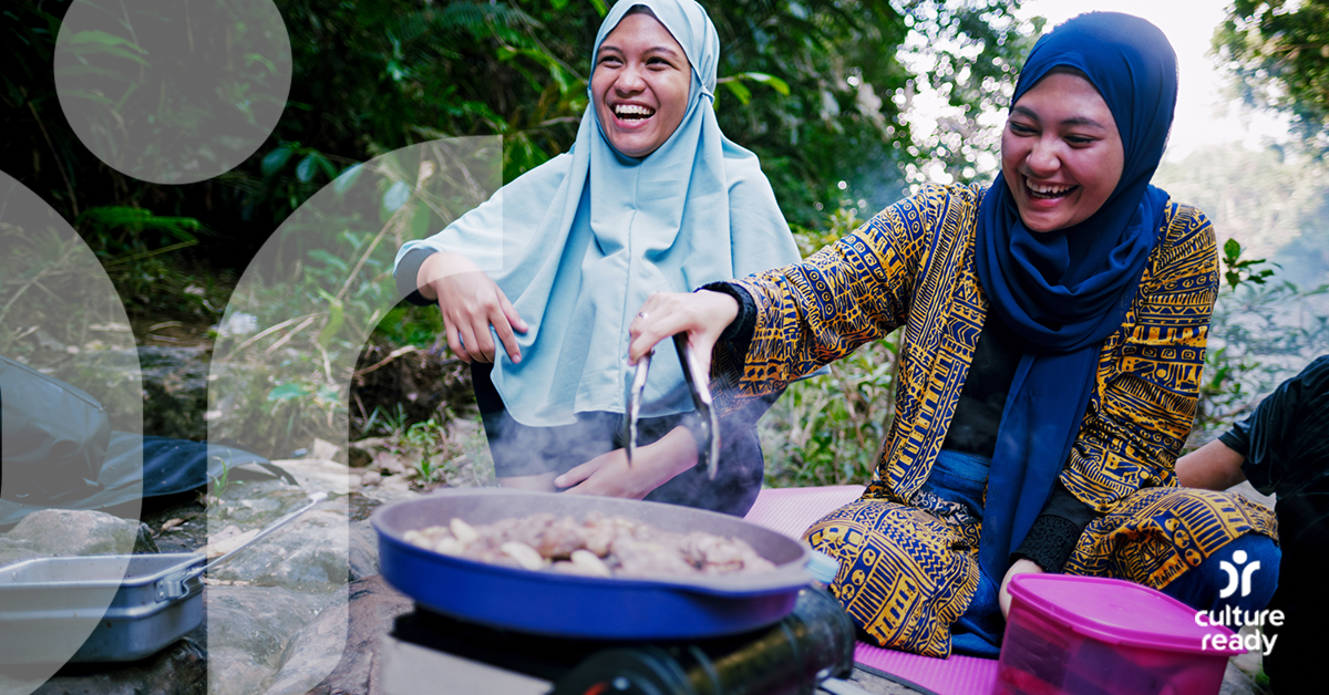 Two women with blue head coverings are laughing while cooking on a skillet outside