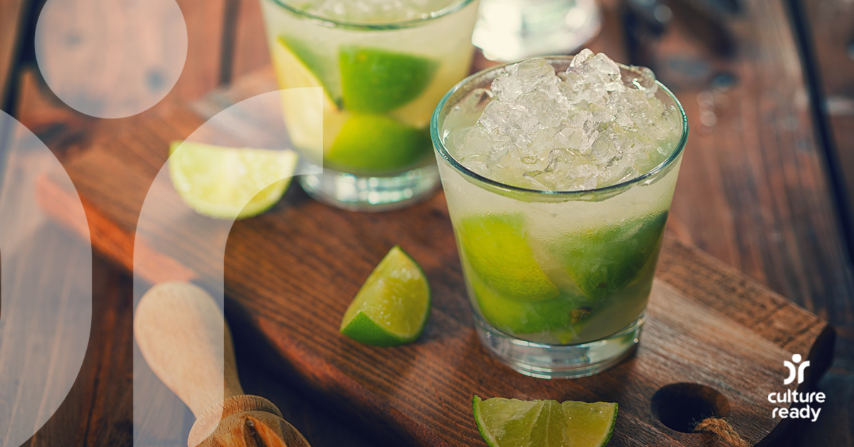 Two cocktail glasses filled with ice, cachaca, and limes sit on a wooden counter with three more slices of lime