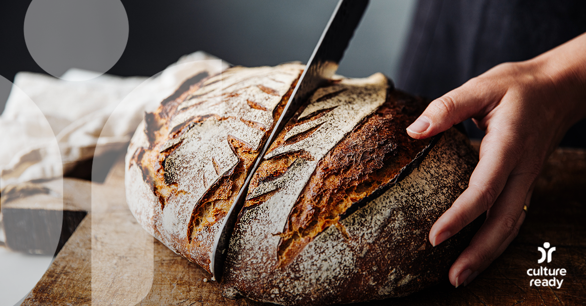 A close up shot of a loaf of traditional german bread. A man holds the loaf with one hand and uses a large knife to cut the loaf in half with the other hand.