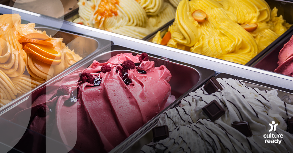 A close up shot of a gelato case shows orange, pink, yellow and white gelato ready to be scooped. The white gelato has chocolate drizzle on top of it.