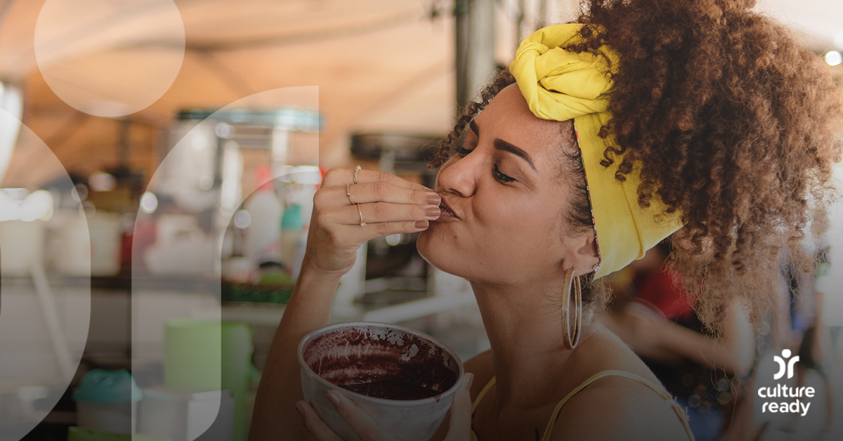 A woman with curly brown hair and a yellow hair scarf is eating a bowl of açai na tigela with a happy expression.