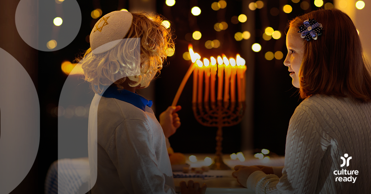 A young boy and girl wearing white with blue accents light a menorah with a night sky background