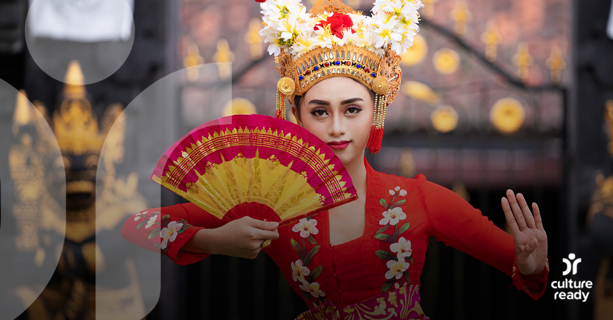A woman in traditional Indonesian makeup wearing a red dress, gold head piece, and carrying a red and yellow fan, does a traditional dance