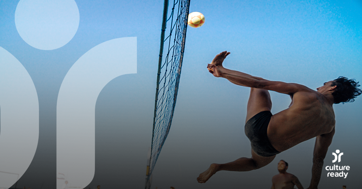 A man serves a footvolley ball over a net while his team mate intently watches in the background