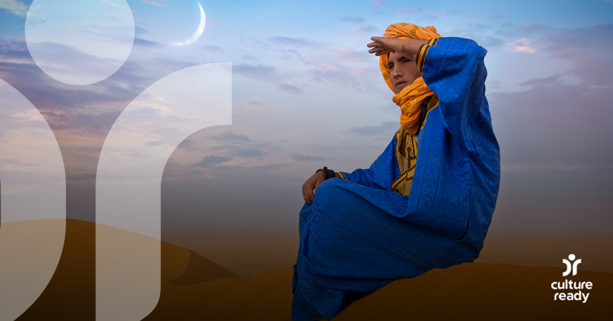 A Berber man in a desert setting sits on a sand dune looking into the horizon wearing a blue tunic and yellow head covering 