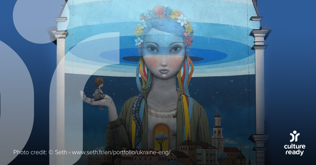 A primarily blue mural on the side of a Kyiv building shows a girl in a flower crown above the city holding a house
