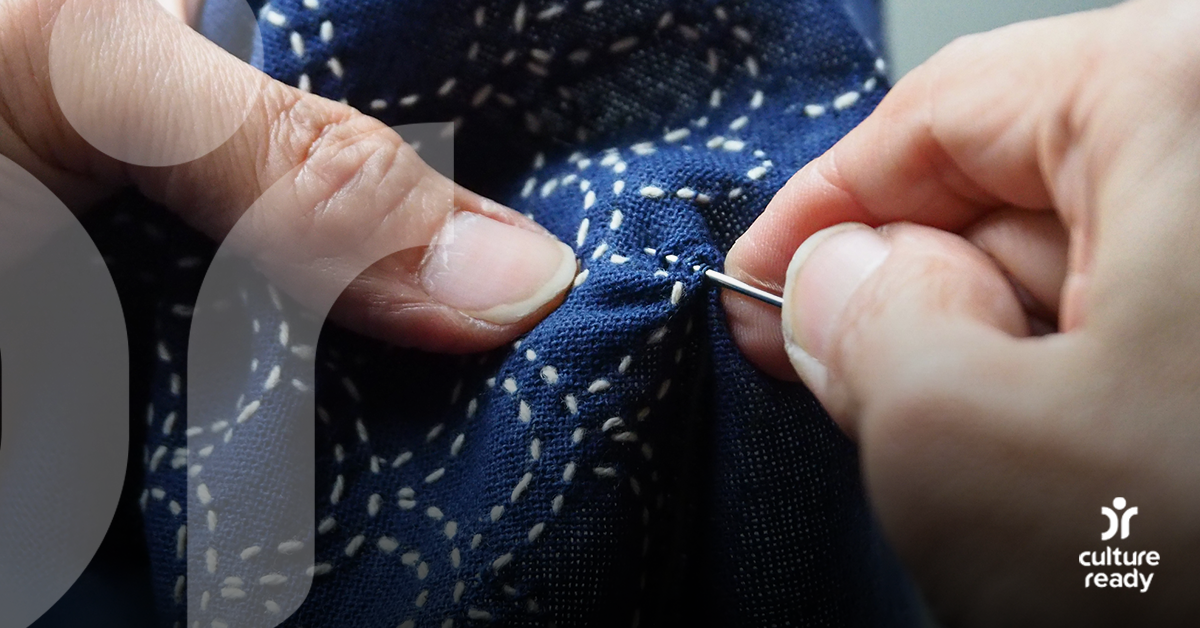 A pair of hands use a needle to thread a pattern with white thread into blue fabric