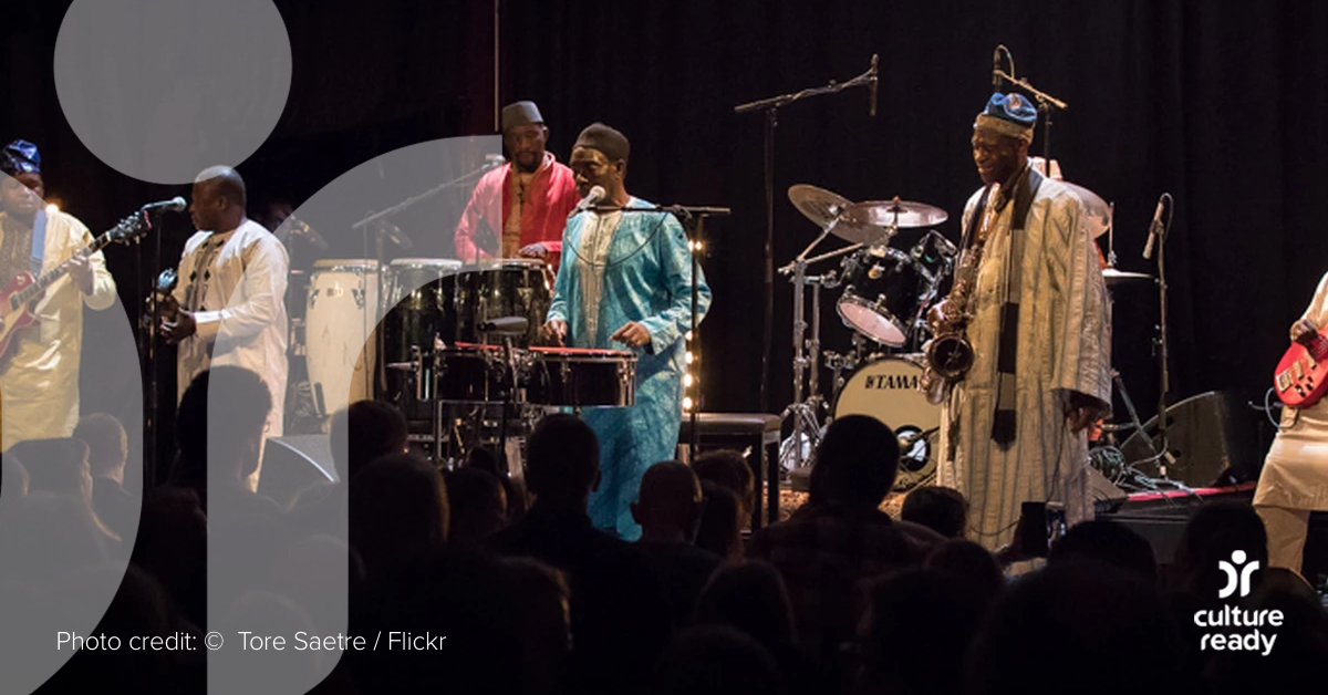Five members of Orchestra Baobab wearing traditional Senegalese clothing play instruments on stage