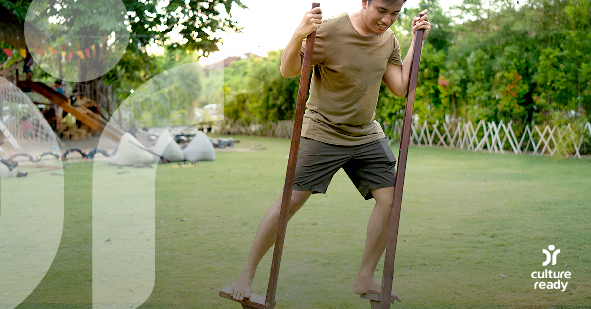 A man wearing green shorts and a green t-shirt practices using bamboo stilts in a field