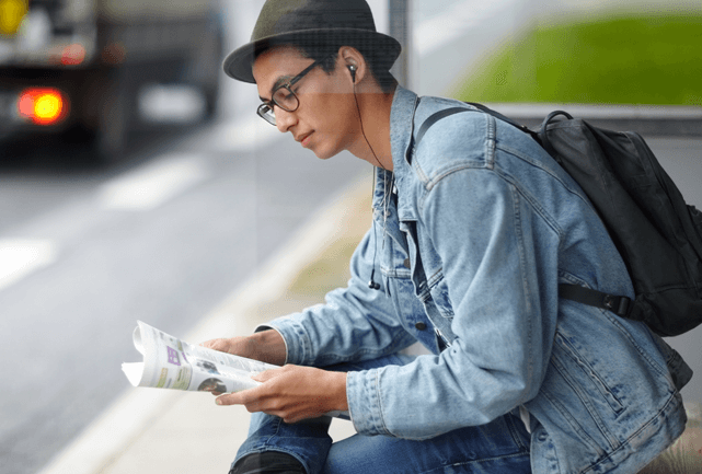 Young man in a hat, sitting at bus stop reading a newspaper.