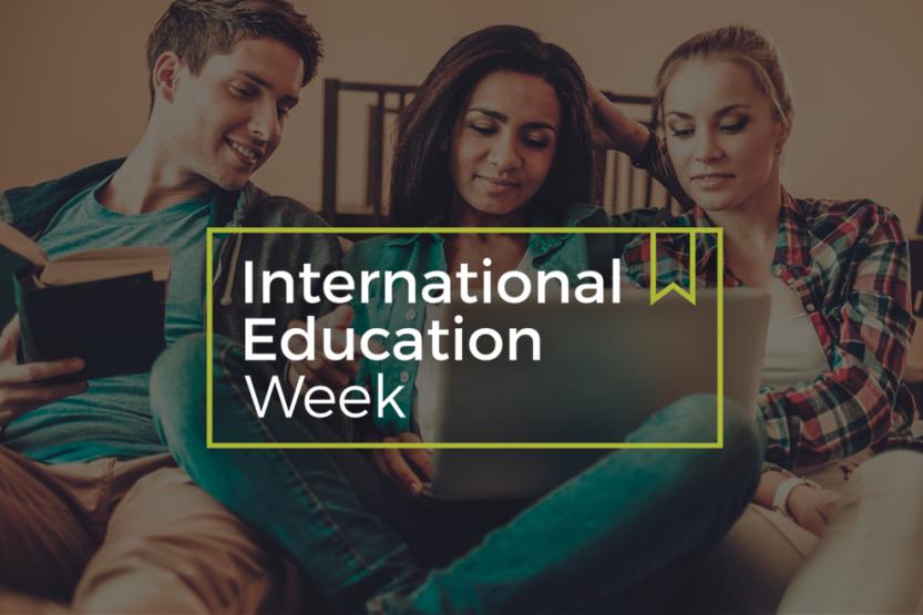 Three people sitting together looking at a person's laptop, with a banner saying "International Education Week" over top