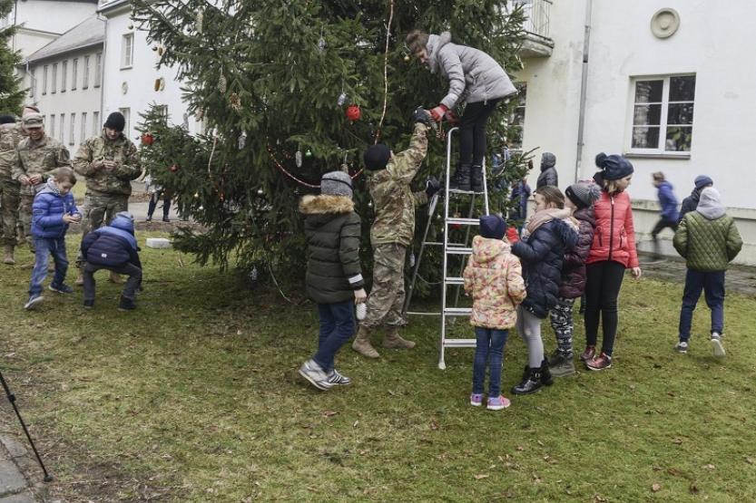 Children and soldiers gathered around a pine tree. A soldier is handing decorations to a woman on a ladder