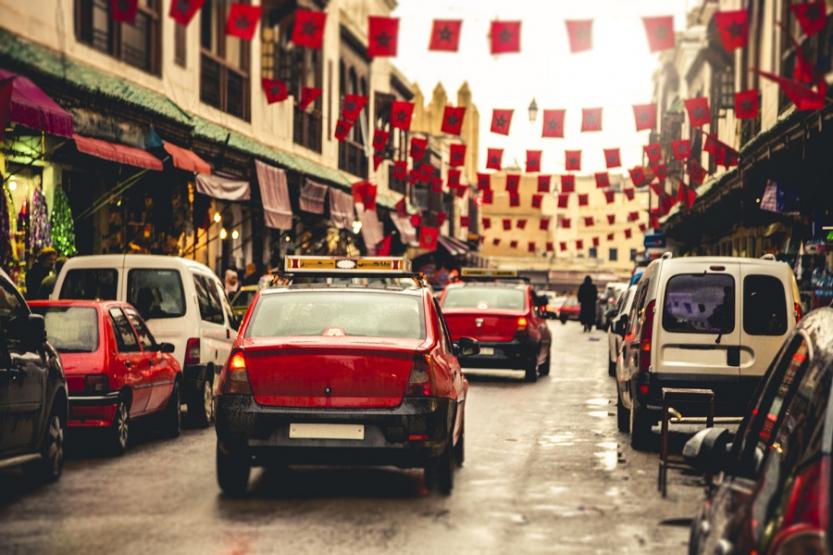 Red taxis in Fez, Morocco, driving down a street with cars parked on either side. Banners of the Moroccan flag hang between buildings.