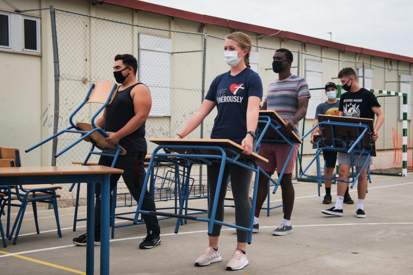 Four people carrying desks and chairs while wearing face masks.