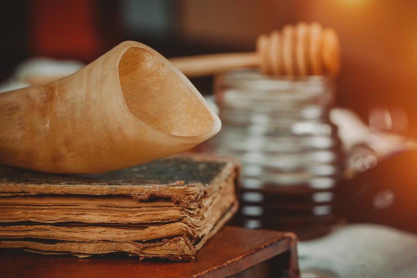 A close-up shot of a shofar, or ram's horn, sitting on top of a worn book. In the background is a jar of honey with a wooden honey dipper on top.