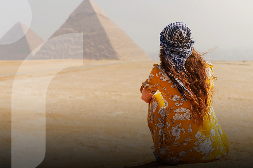 A woman wearing a bright orange dress and a blue checked headscarf facing away from the camera. She is looking at Egyptian pyramids in the distance.