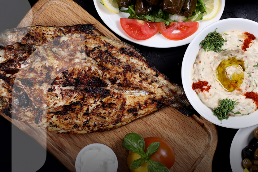 A large piece of grilled fish on a cutting board with vegetables, hummus, and olives on the side