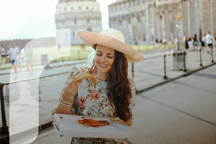 A woman in a floral dress and straw hat eats a slice of pizza in front of a Roman cityscape