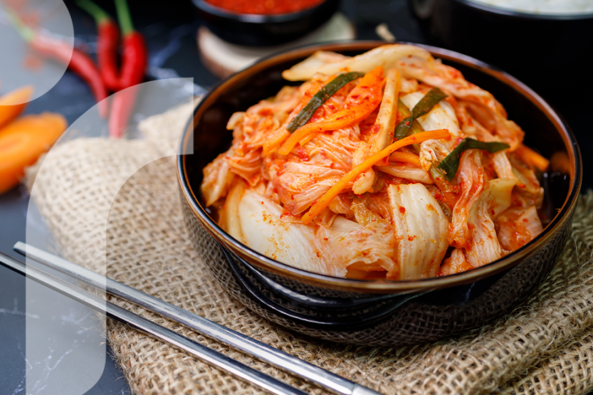Kimchi in a black bowl is placed on top of a burlap table covering near silver metal chopsticks