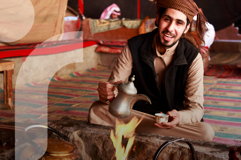 A Bedouin man in traditional clothing pours a cup of coffee in front of a fire